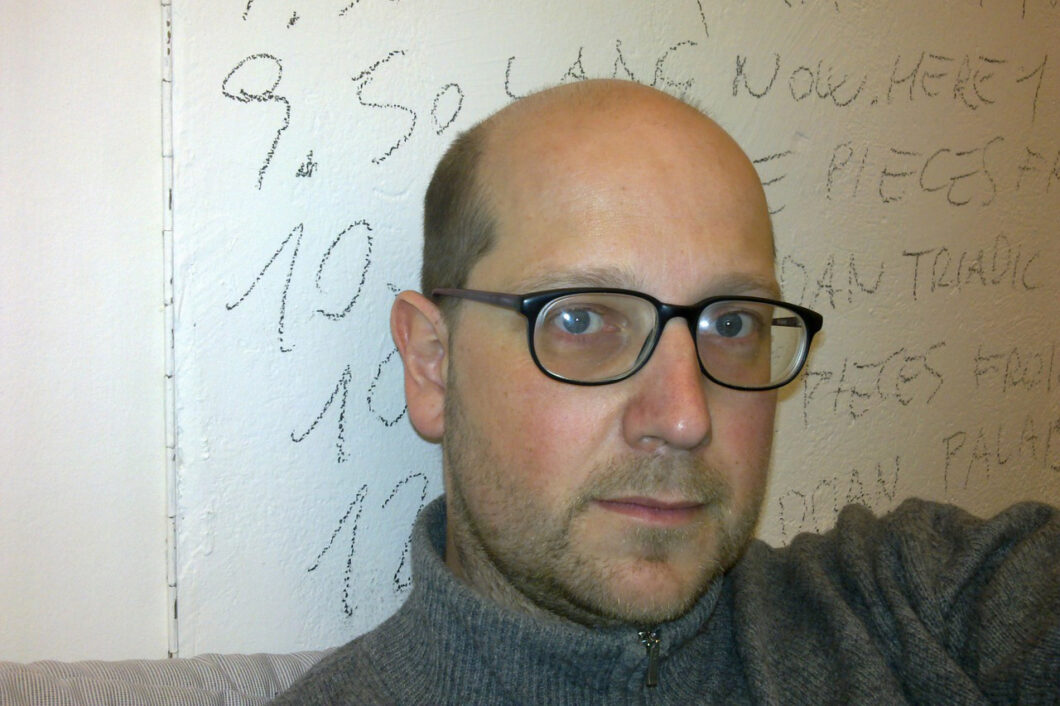 Ein Mann mit Glatze und schwarz umrandeter Brille sitzt vor einer Wand, an der Zahlen geschrieben stehen. Es ist Komponist Klaus Lang.