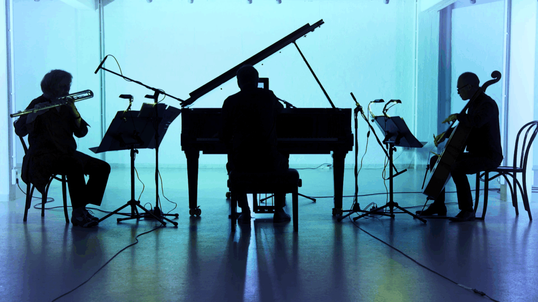 Es ist der Schattenriss vor hellblauem Licht von einer Person mit Querflöte, einer Person am Klavier und einer Person mit Cello; es ist das Ensemble L'ART POUR L'ART.