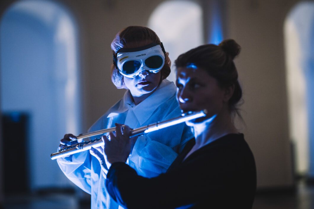Eine Frau spielt Querflöte, eine Person in einem weißen Kittel mit einer weißen Taucherbrille schaut sie an, beide werden von blauem Licht angestrahlt; es sind Viktoriia Vitrenko und Eva Ludwig.