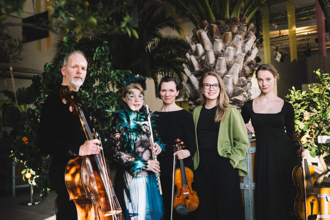 Ein Cellist, eine Flötistin, eine Geigerin, eine Frau in grüner Jacke und eine Bratschistin stehen mit ihren Instrumenten in einem Raum vor Palmen; es ist das Ensemble musica assoluta.