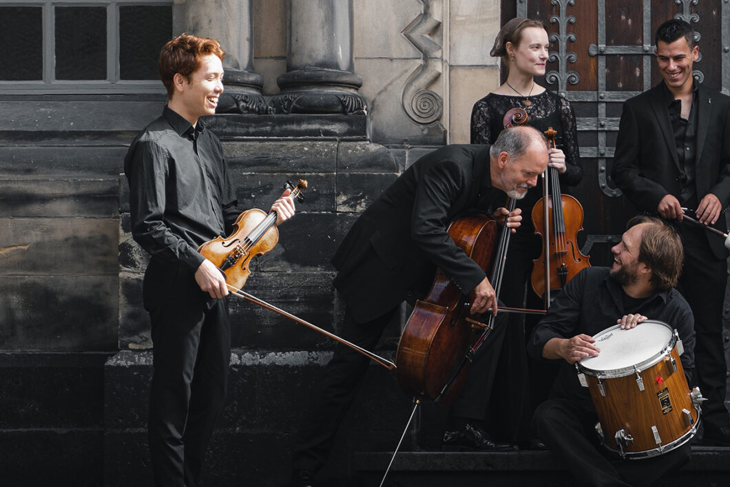 Musiker*innen mit Geige, Cello, Trommel und Bratsche stehen vor einer Steintreppe und unterhalten sich fröhlich. Es sind Musiker*innen des hannoverschen Ensembles musica assoluta.