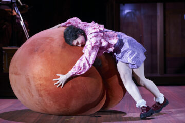 Eine Person in einem Kostüm aus rosa Bluse und lila Rock umarmt einen riesigen Pfirsich. Es ist eine Szene aus "Was ihr wollt" nach William Shakespeare.